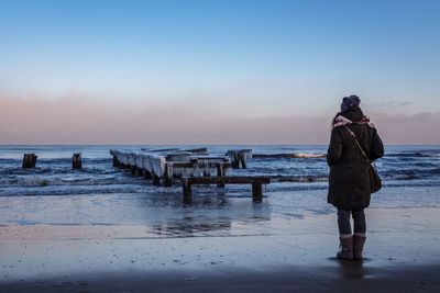 Woman looking at frozen wooden posts on sea shore against sky