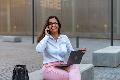 Young woman using smart phone while sitting on laptop