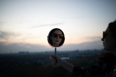Reflection of woman wearing sunglasses in mirror against sky during sunset