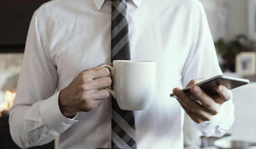 Midsection of businessman holding coffee cup using smart phone while standing in office