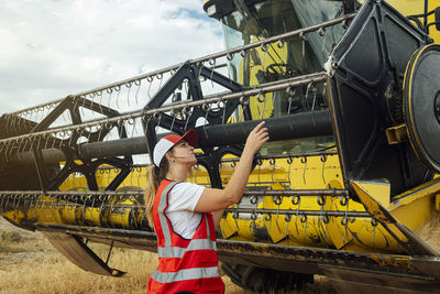 Side view of focused female farmer examining metal reel of industrial combine harvester while working in agricultural field of countryside