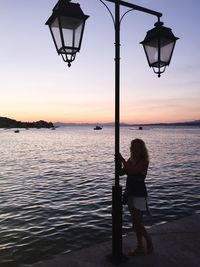 Full length of woman standing by lamp post while standing on pier against sky during sunset