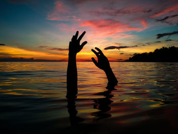 Silhouette cropped hands of man in lake against cloudy sky at sunset