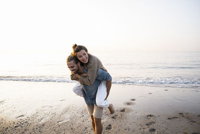 Happy man giving piggyback to girlfriend while walking on shore at beach against clear sky during sunset