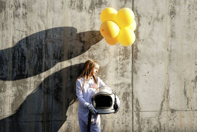 Girl holding balloon while standing against wall on sunny day