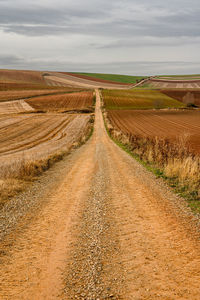 Dirt road passing through field against sky