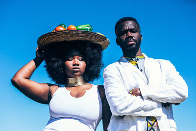 Trendy black woman in white dress with metal plate with vegetables on head standing near confident man with crossed arms against cloudless blue sky