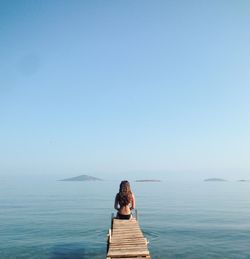 Rear view of woman sitting on pier over sea against clear sky