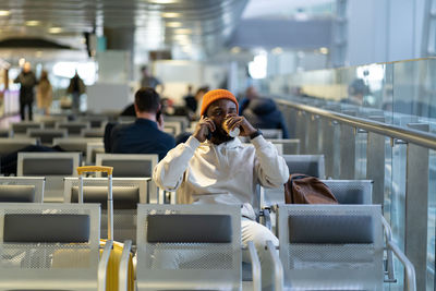 African traveler man drinking coffee waiting for flying at airport terminal, talking on cellphone.