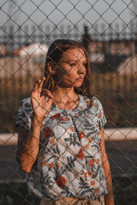 Thoughtful young woman looking through chainlink fence at sunset