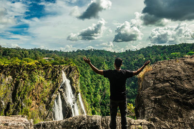 Rear view of man standing on rock against waterfall and sky