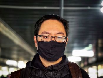 Portrait of man wearing eyeglasses and black face mask against ceiling, wall and lights.