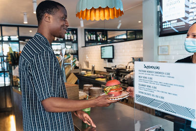 Side view of content black male with tasty burger on plate looking forward against signboard in restaurant