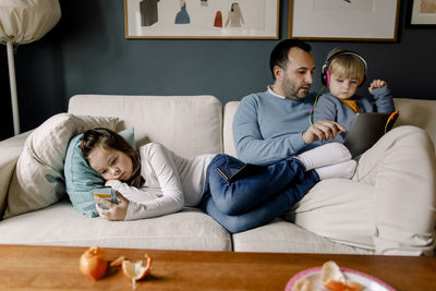 Girl using mobile phone while father showing digital tablet to sister on couch in living room at home