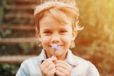 Portrait happy dishevelled long blond hair preschool kid boy holding delicate pale lilac wildflower