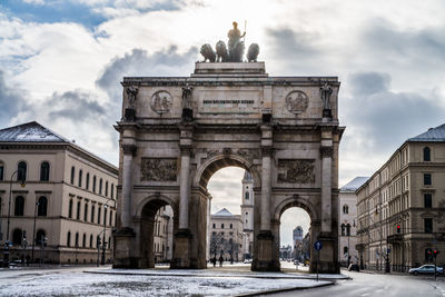 Low angle view of siegestor with buildings against cloudy sky