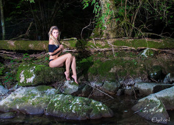 Full length of beautiful woman sitting on rock at lakeshore in forest