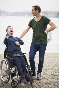 Male caretaker standing with happy disabled man on wheelchair by lake