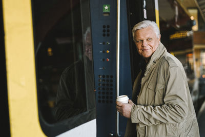 Side view of happy businessman holding disposable cup while boarding in train at railroad station