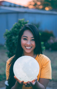 Portrait of smiling young woman holding illuminated ball on field during sunset