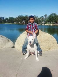 Boy sitting leaning on rock with pit bull terrier against lake