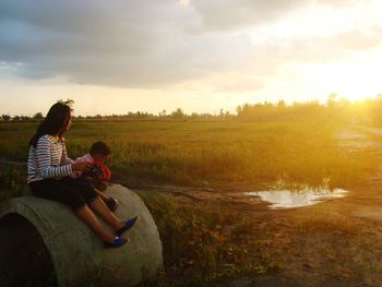 Full length of woman sitting on son on stone at field during sunset