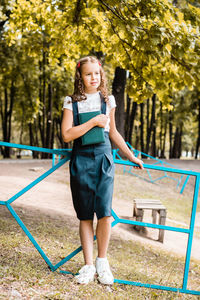 A female student in a school uniform with a book stands in the park on a warm day. vertical view