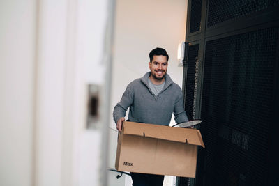 Smiling man carrying cardboard box while walking by metal grate in house