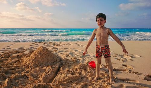 Full length of shirtless boy standing at beach against sky