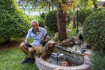 Garden. a mature man by the small fountain has a break while working in the tools.