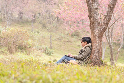 Young woman using laptop while sitting by tree on field