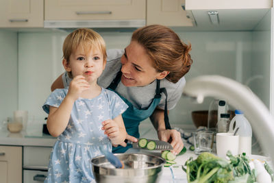 Funny toddler daughter helps mom cook in the home kitchen and eats vegetables.