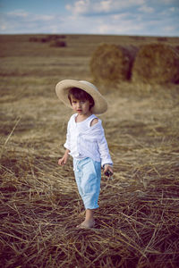 Boy a child in a straw hat and blue pants stands in a mowed field with stacks in the summer