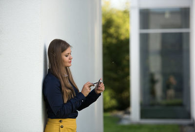 Side view of smiling young woman using mobile phone by concrete wall