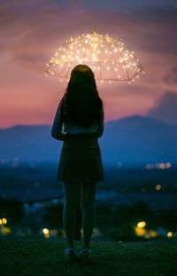 Rear view of woman with illuminated umbrella standing on field against sky during sunset