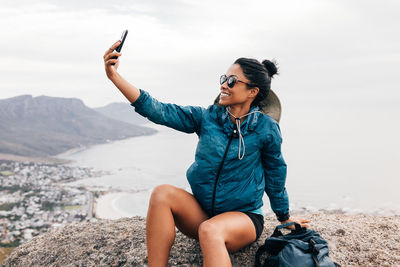 Smiling woman taking selfie while sitting on rock against sea
