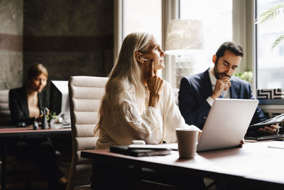 Mature businesswoman wearing in-ear headphones at laptop by businessman at desk in office