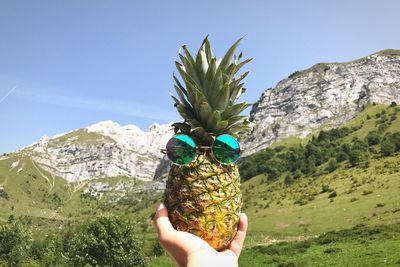 Cropped image of hand holding pineapple with sunglasses against mountains