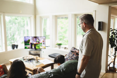 Father changing channels of television through remote control while standing in living room at home