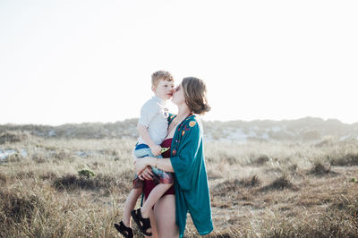Mother kissing son while standing on grassy field against clear sky during sunny day