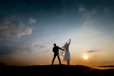 Rear view of bride and groom standing on field against sky during sunset