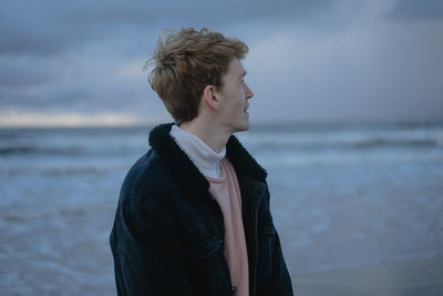 A guy with brown hair enjoying the seascape, in a turtleneck and a warm denim black jacket