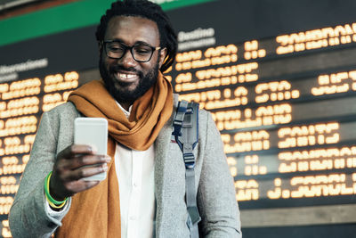 Smiling man using mobile phone while standing airport departure board