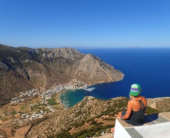 Woman sitting on rock by sea against clear blue sky