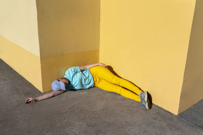 Mid adult woman lying on footpath by yellow wall during sunny day