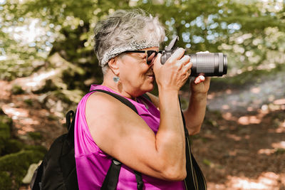 Side view of elderly female tourist with backpack shooting nature while spending sunny summer day in park