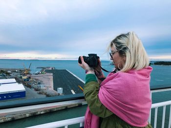 Side view of woman photographing sea against cloudy sky