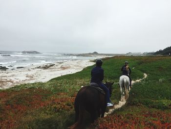 Rear view of people riding horses at sea shore against sky