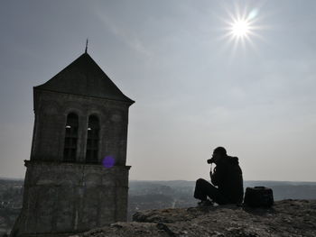 Silhouette of man photographing sitting on rock against sky