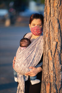 Portrait of mother wearing carrying daughter by tree trunk